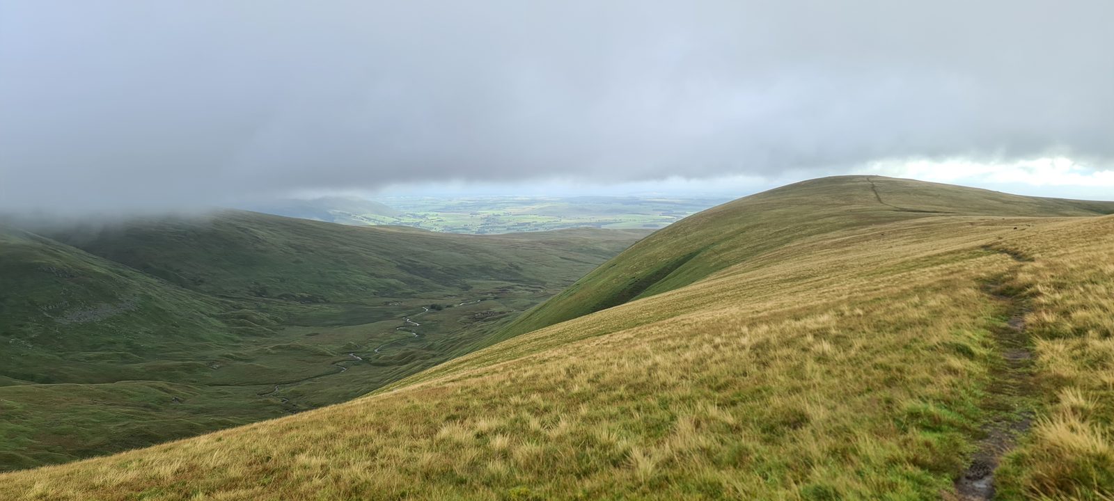 A grassy fellside falling away to a deep valley, with a worn path running along the ridge to the right and cloud pressing down on the tops