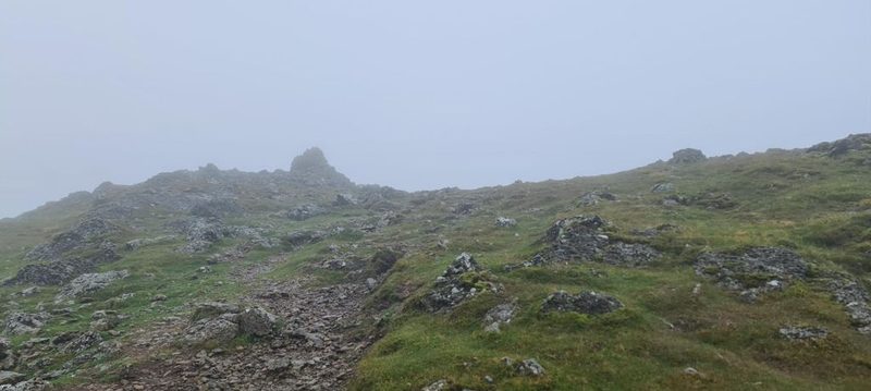 A stony path climbing through mist towards a rocky top, grass and lichen-covered boulders to either side and visibility closing in