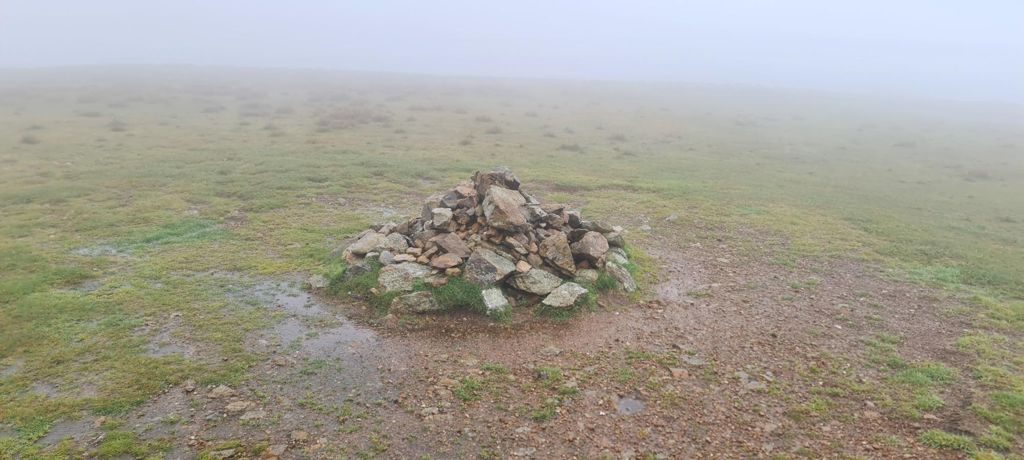 A small stone cairn on a flat, featureless grassy plateau, with thick mist closing in on every side