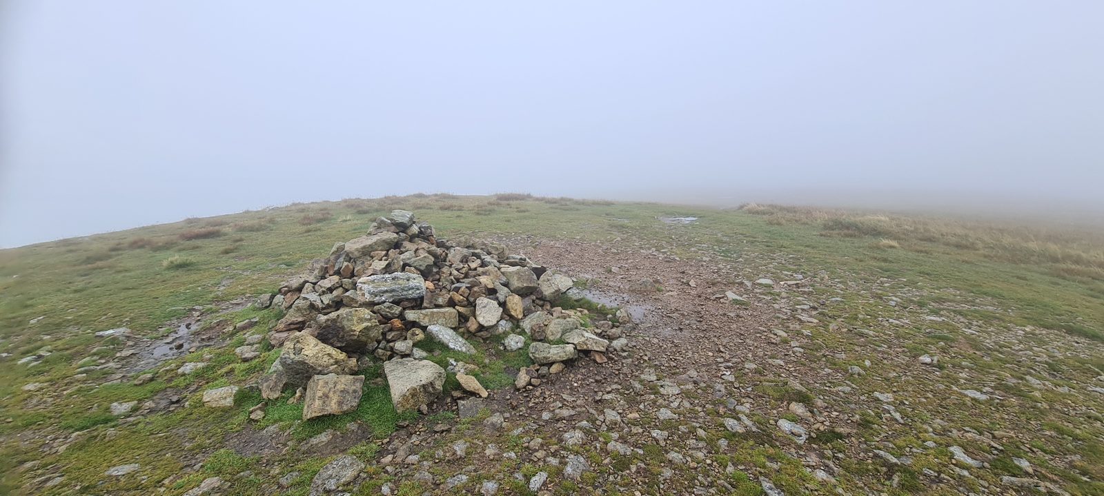 A stone cairn beside a stony path on a broad summit, everything beyond it lost in thick grey mist