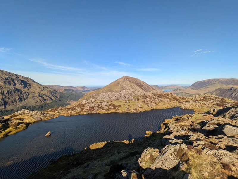 Innominate Tarn on the summit plateau of Haystacks, dark and wind-ruffled among rock and heather, with a shapely fell rising beyond