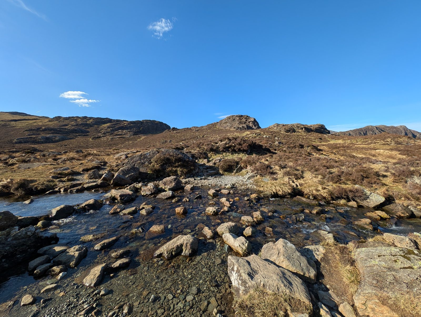 A shallow beck spreading over bare rock slabs and boulders, with a rough path climbing beyond it towards a rocky knott