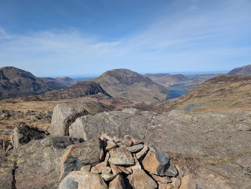 A cairn on bare rock slabs looking out over a rocky top to Crummock Water and Buttermere lying in their valleys far below