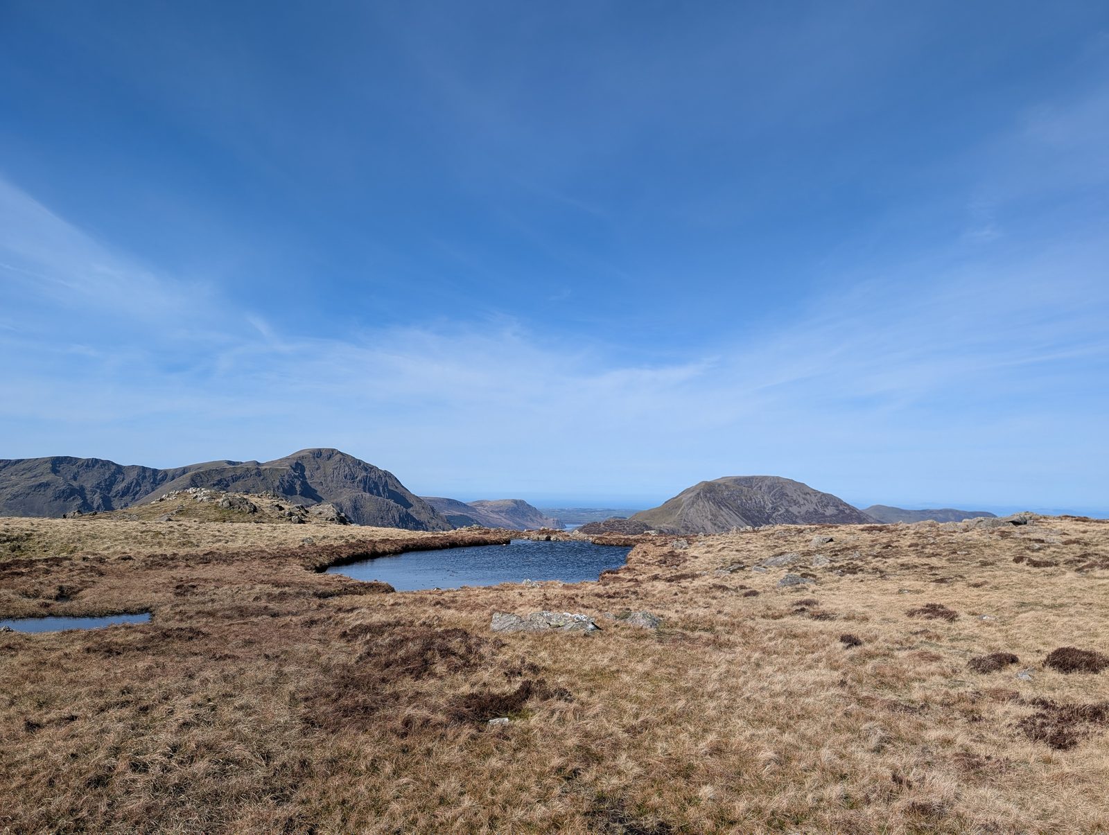A small dark tarn set in a bare, tussocky plateau, with crag-topped fells on the left and a rounded top on the right against a pale blue sky