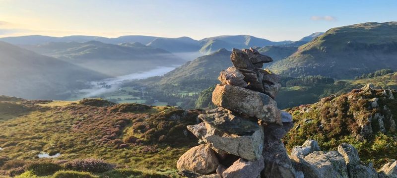 A crag-topped fell rising beyond a drystone wall in early light, with the moon still pale in the blue sky above it