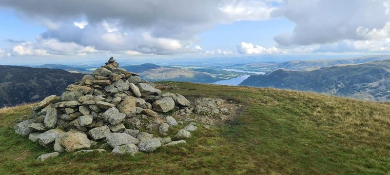 A large stone cairn on the broad grassy summit of Birkhouse Moor, with Ullswater and the low country stretching away beyond