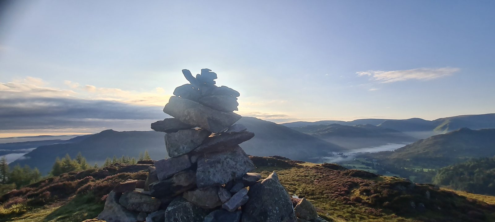 A crag-topped fell rising beyond a drystone wall in early light, with the moon still pale in the blue sky above it