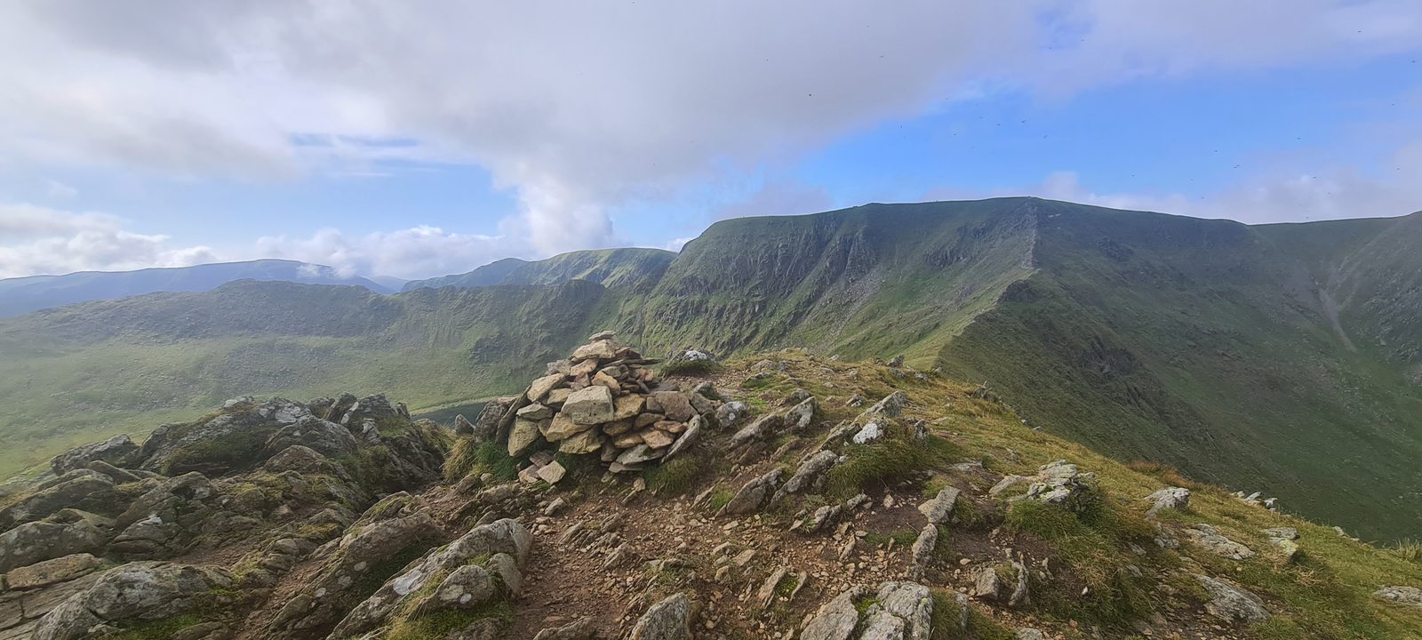 A cairn on a stony ridge crest looking across to a great crag-rimmed hollow, with a path climbing the skyline beyond