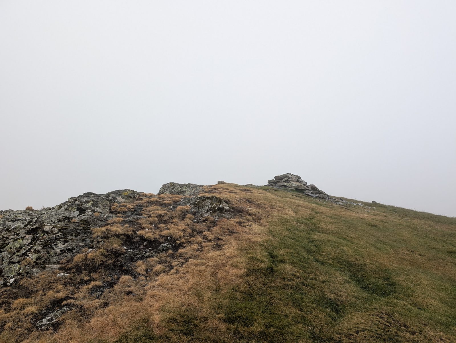 A small cairn on a rocky, grassy summit ridge with thick white cloud closing in behind it and dead bleached grass in the foreground