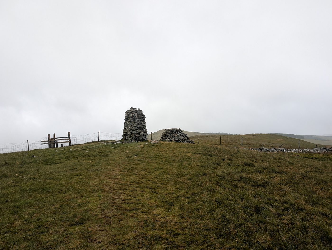 The tall summit cairn on Broom Fell beside a fence and gate, the fells beyond lost in low cloud