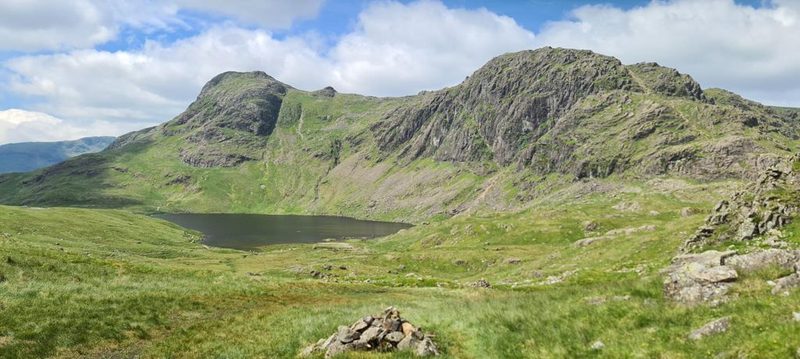 Stickle Tarn from above, dark under the sun, with the craggy Langdale Pikes rising steeply behind it and a small cairn on the grass in front