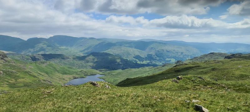 A high grassy shoulder looking down over a small tarn to a green valley, with ridge after ridge of fells running to the horizon