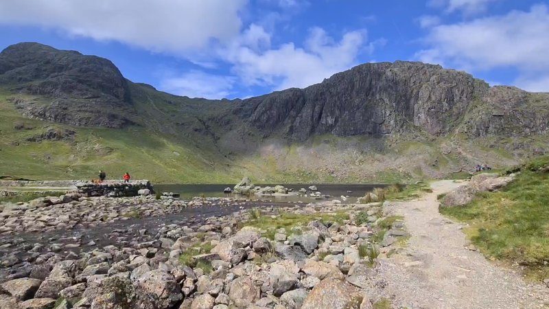 Stickle Tarn below the great crag of Pavey Ark, with walkers crossing the stone outflow and a boulder-strewn path in the foreground