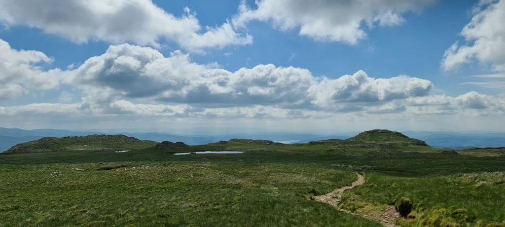 A broad, gently rolling grassy plateau with small pools and low rocky knolls, a thin path running away under a sky of scattered cumulus