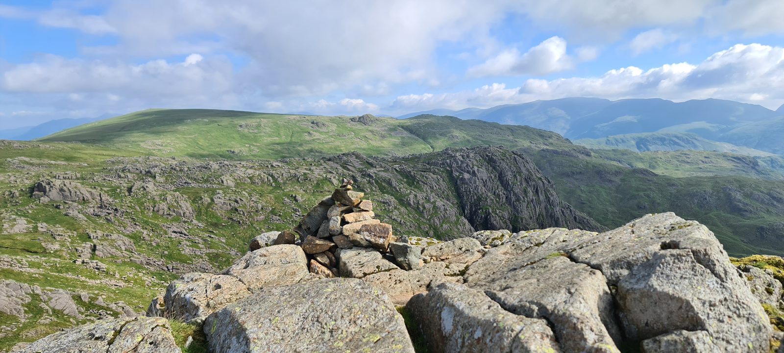 A cairn on a rocky top with a deep craggy gully falling away beyond it, and high fells ranged along the skyline