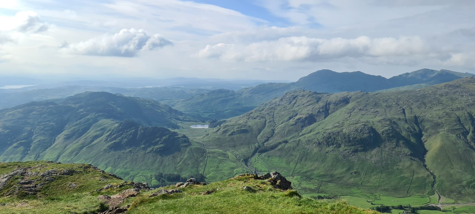 Looking down the length of a green valley from a grassy summit, with a small tarn far below and range after range of fells on either side