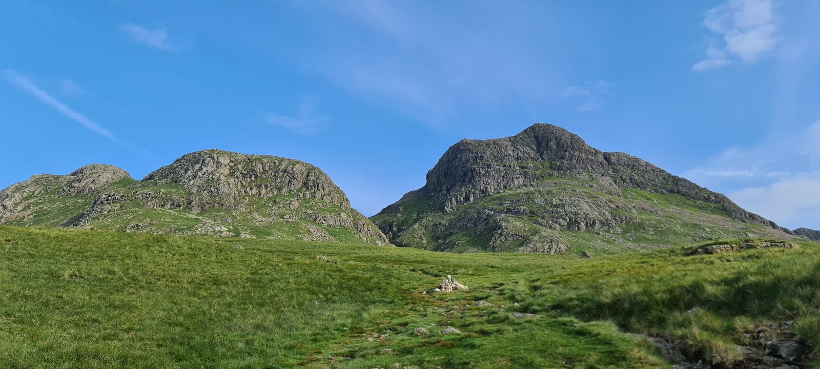 The rocky crest of the Langdale Pikes seen from below on the descent, with a grassy path leading down through the foreground