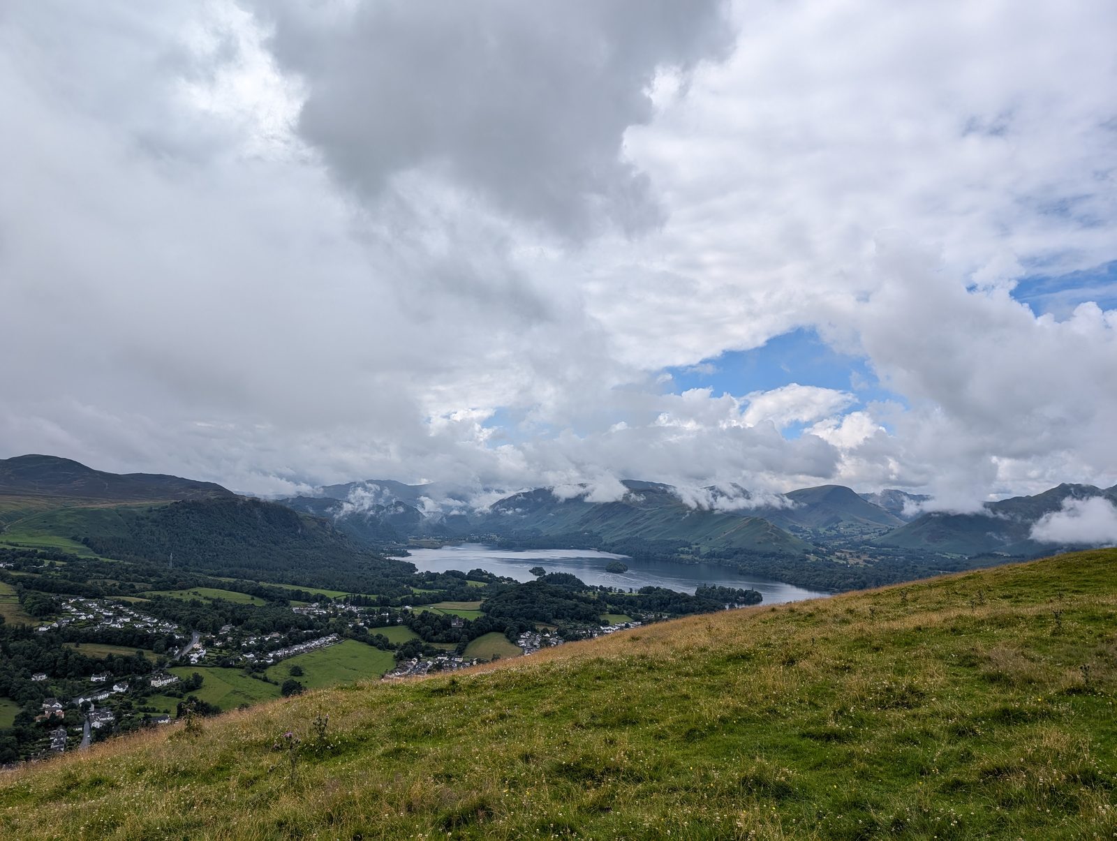 Derwentwater and the rooftops of Keswick from the grassy south side of Latrigg, with cloud lying along the north-western fells beyond the lake