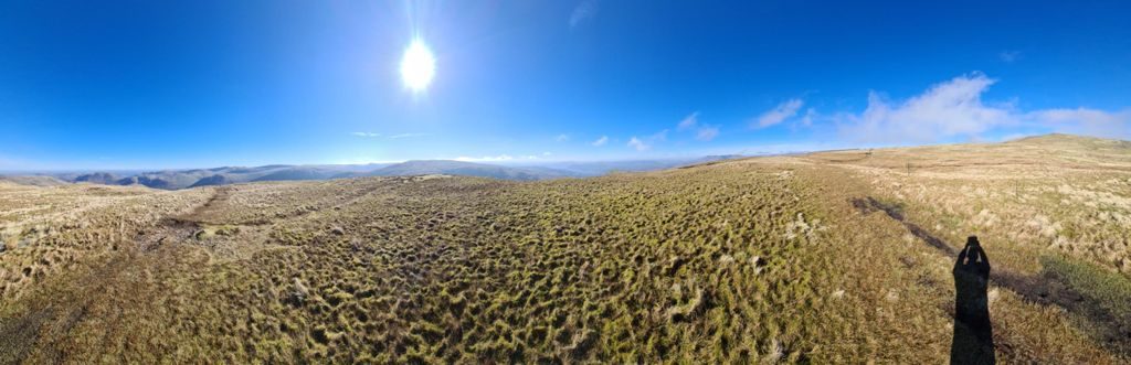 A wide view across a broad, tussocky plateau of pale moor grass under a high sun, with ranks of distant fells along the horizon and the photographer's shadow cast across the grass