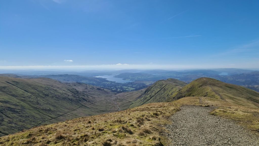 A broad stony path curving down a grassy ridge, with a deep valley falling away on the left and Windermere shining in the distance