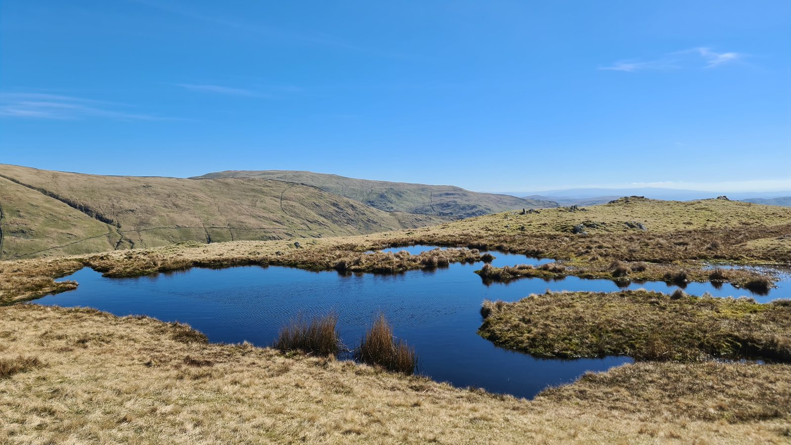 A small dark tarn set among rough tussocks on a broad grassy ridge, with a long fellside and a stony escarpment rising beyond under a clear blue sky