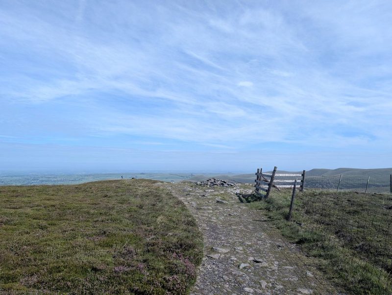 A stony path running along a broad heathery ridge to a small cairn, with a wooden ladder stile over the fence on the right and the Solway plain beyond