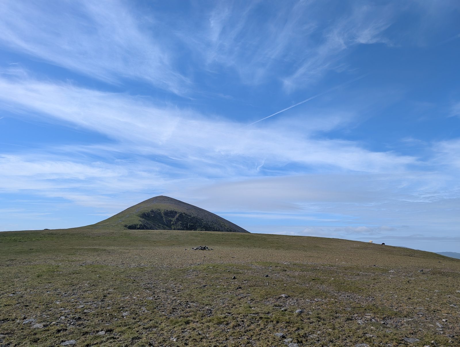 A broad, bare grassy plateau with a low cairn, and beyond it a steep conical fell with thin cloud forming over its top