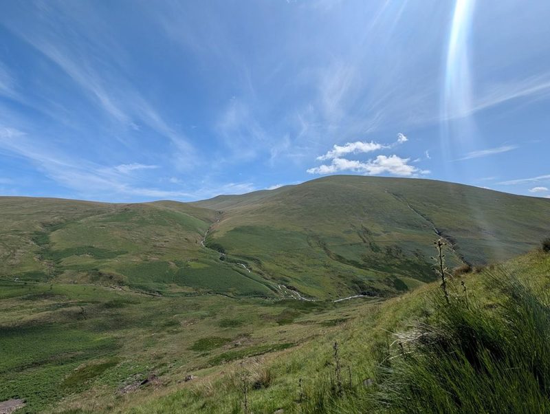 A sun pillar in a blue sky above the smooth green flanks of the fell, with becks threading down the hillside and bracken filling the valley floor