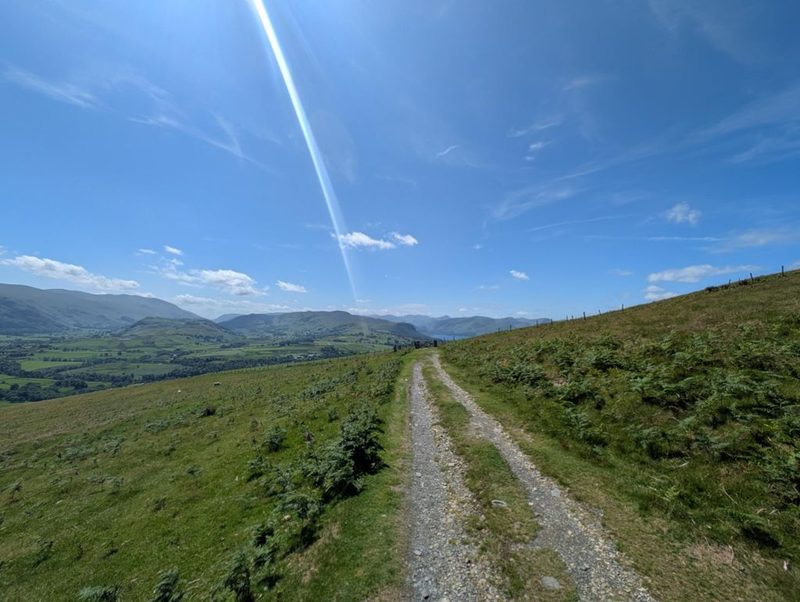 A stony vehicle track running along the open fellside toward distant fells, with bracken on the banks and a wire fence following the ridge above