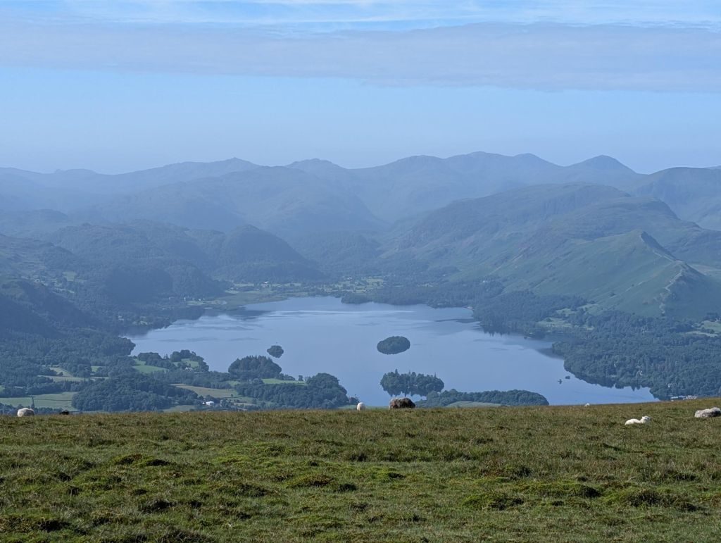 A small stone cairn on a broad grassy shoulder high on the climb, with a long ridge of higher fells filling the skyline across the valley behind