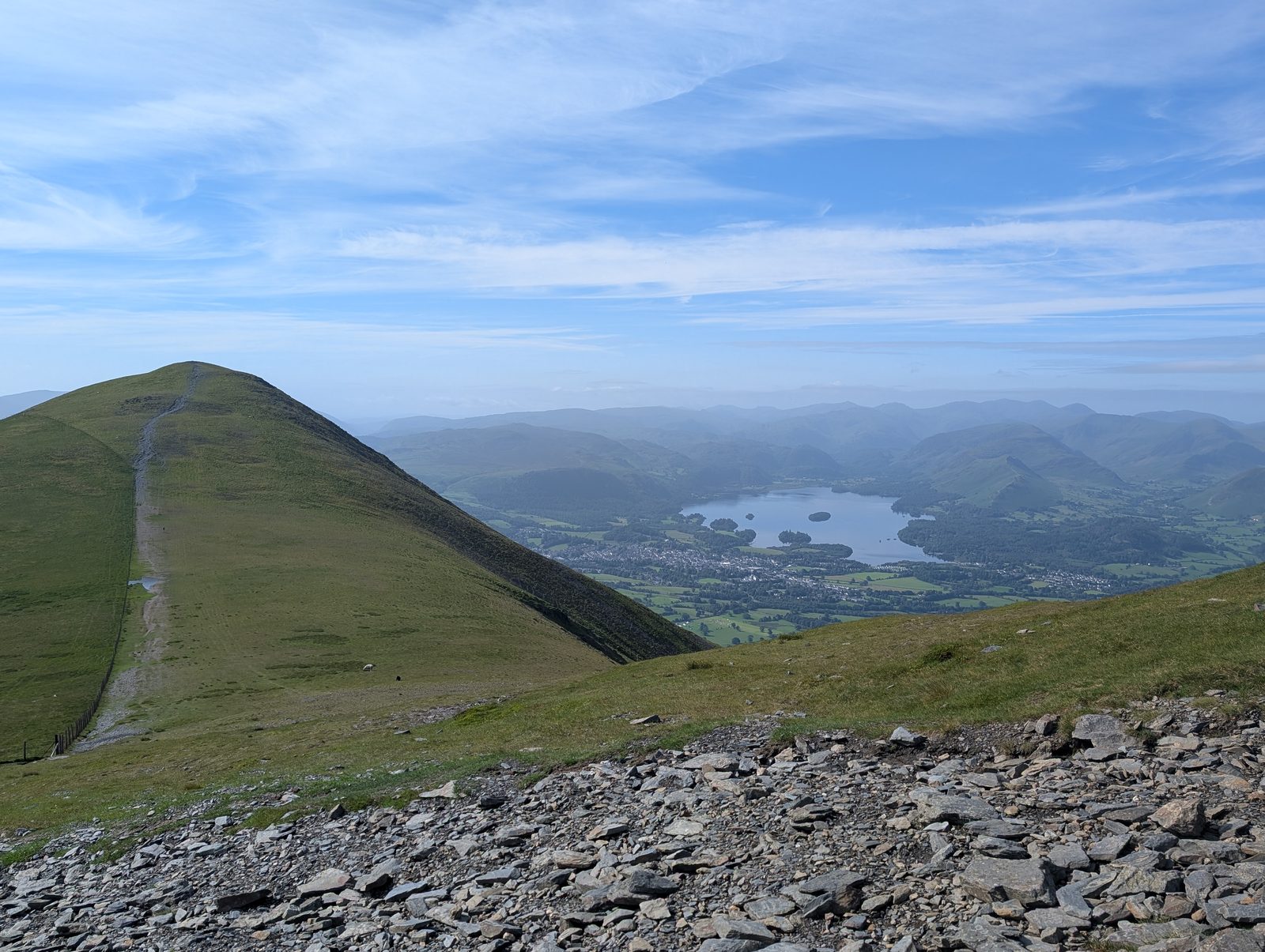 The grassy cone of Skiddaw Little Man ahead with a path running straight up its spine, Derwentwater and Keswick in the valley to the right and slate scree in the foreground