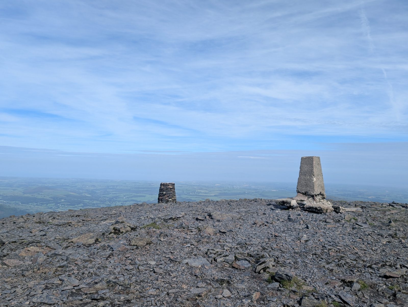 The concrete trig pillar and a stone cairn on Skiddaw's broad stony summit, with the Solway plain stretching flat and hazy to the horizon