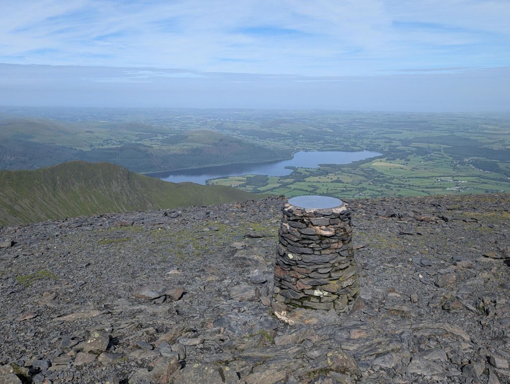 A drum-shaped stone cairn with a metal viewfinder plate on Skiddaw's summit ridge, looking down over Bassenthwaite Lake and the patchwork farmland beyond