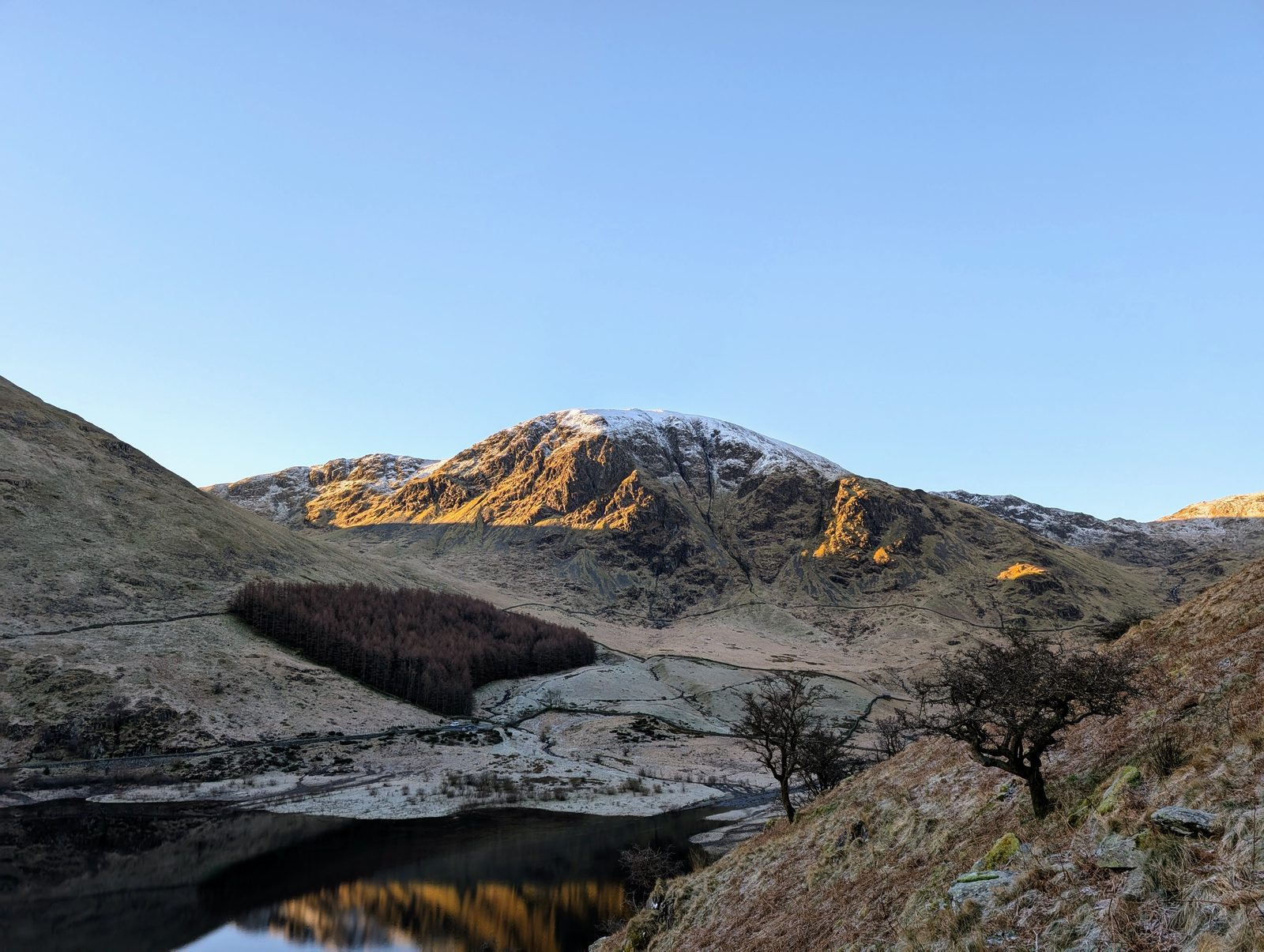 Early sun catching the snow-dusted crags above a still reservoir, with a dark plantation and bare hawthorns on the frosted valley floor