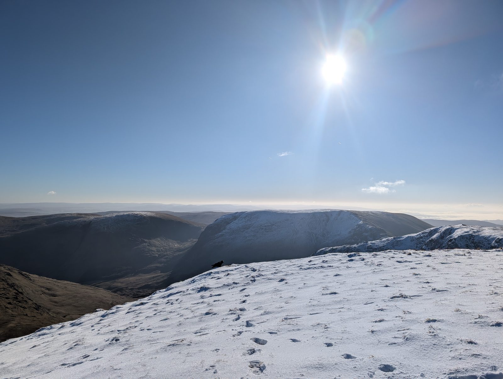 A snow-covered ridge crest with the low sun blazing above it, footprints leading away and a long snowy escarpment beyond
