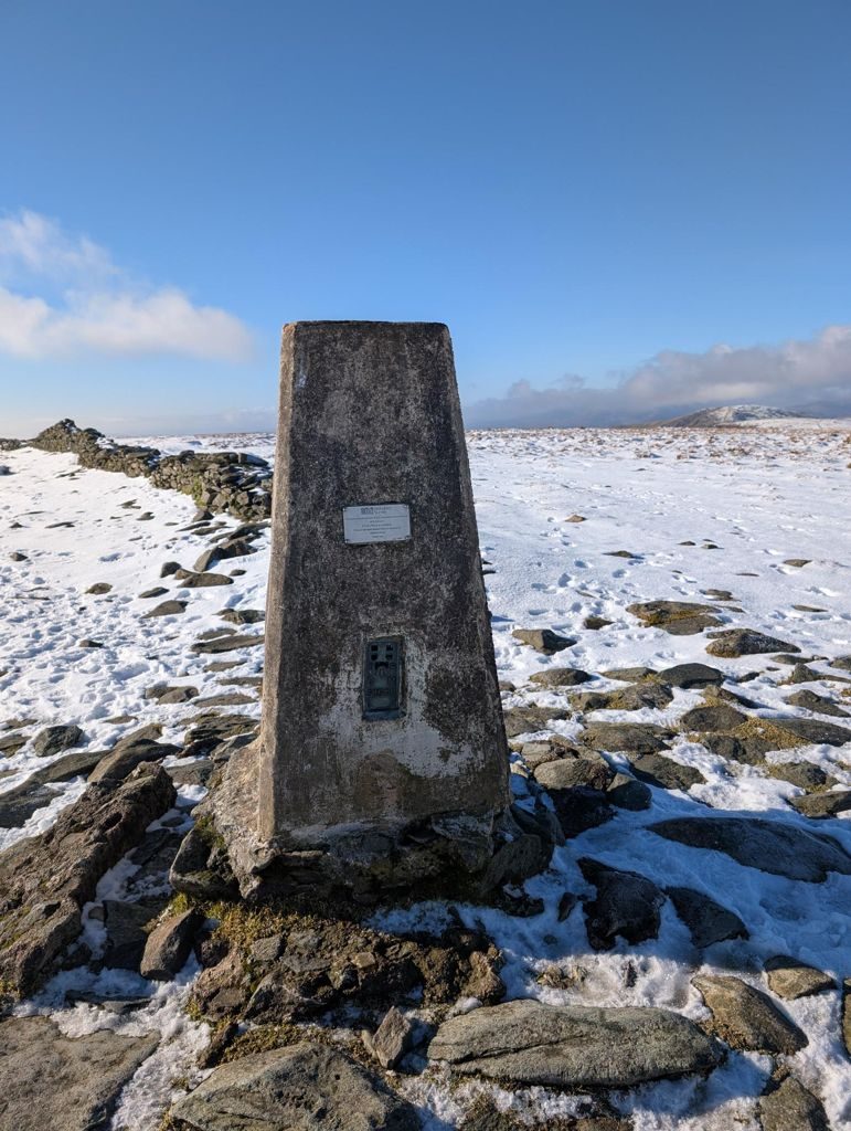 The concrete trig pillar on High Street standing in fresh snow, with a broken stone wall to the left and the plateau running flat to the horizon