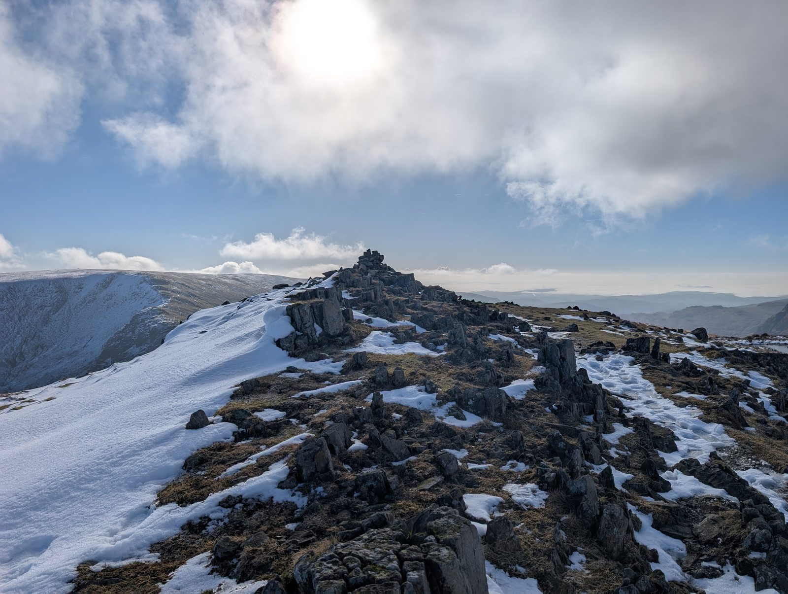 A rocky ridge under fresh snow leading to a small cairn, with a broad snow-covered fellside falling away on the left