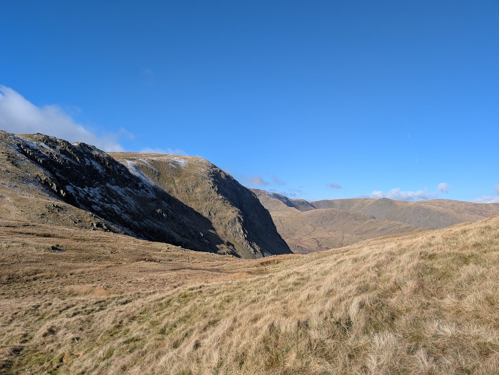 A dark crag streaked with snow above a wide sweep of pale winter grass, with a deep valley and long rounded ridges beyond