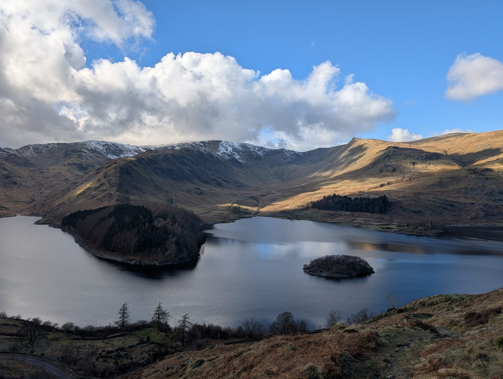Haweswater from above, its wooded promontory and small island dark against the water, with snow-topped fells ranged along the far side
