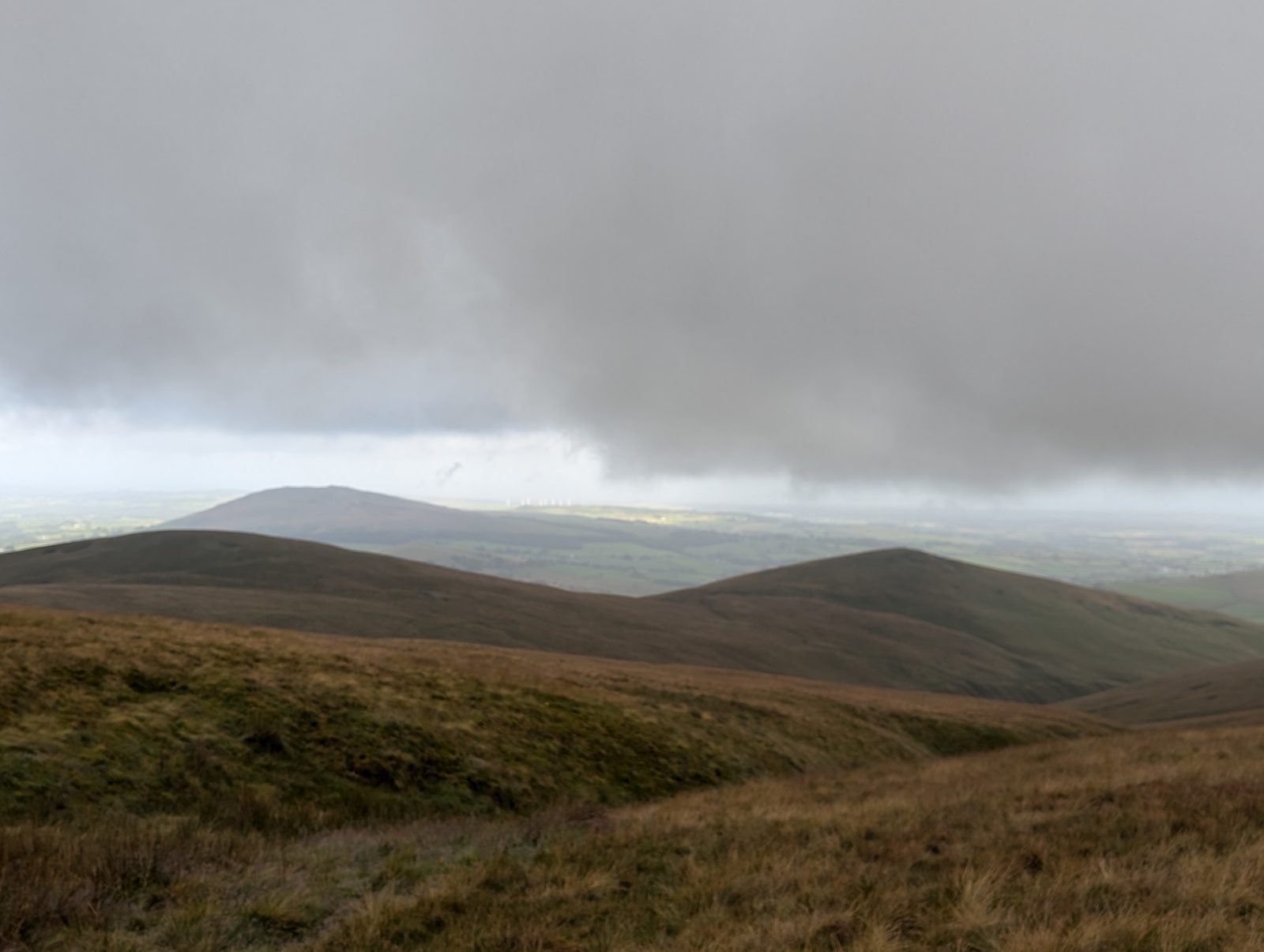 A low bank of rain cloud pressing down over rounded moorland tops, with the flat farmland of the northern plain pale and sunlit beneath it