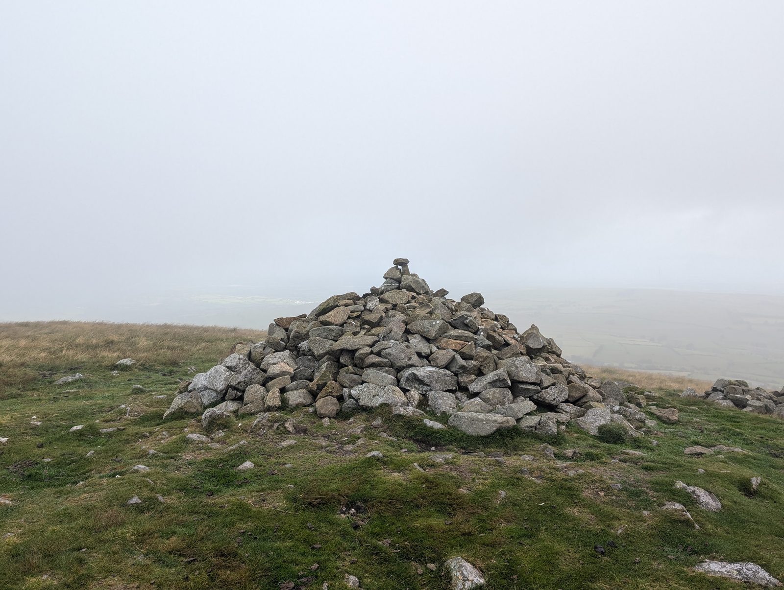 A large stone cairn on a bare grassy summit in thick mist, with the ground falling away into cloud on all sides
