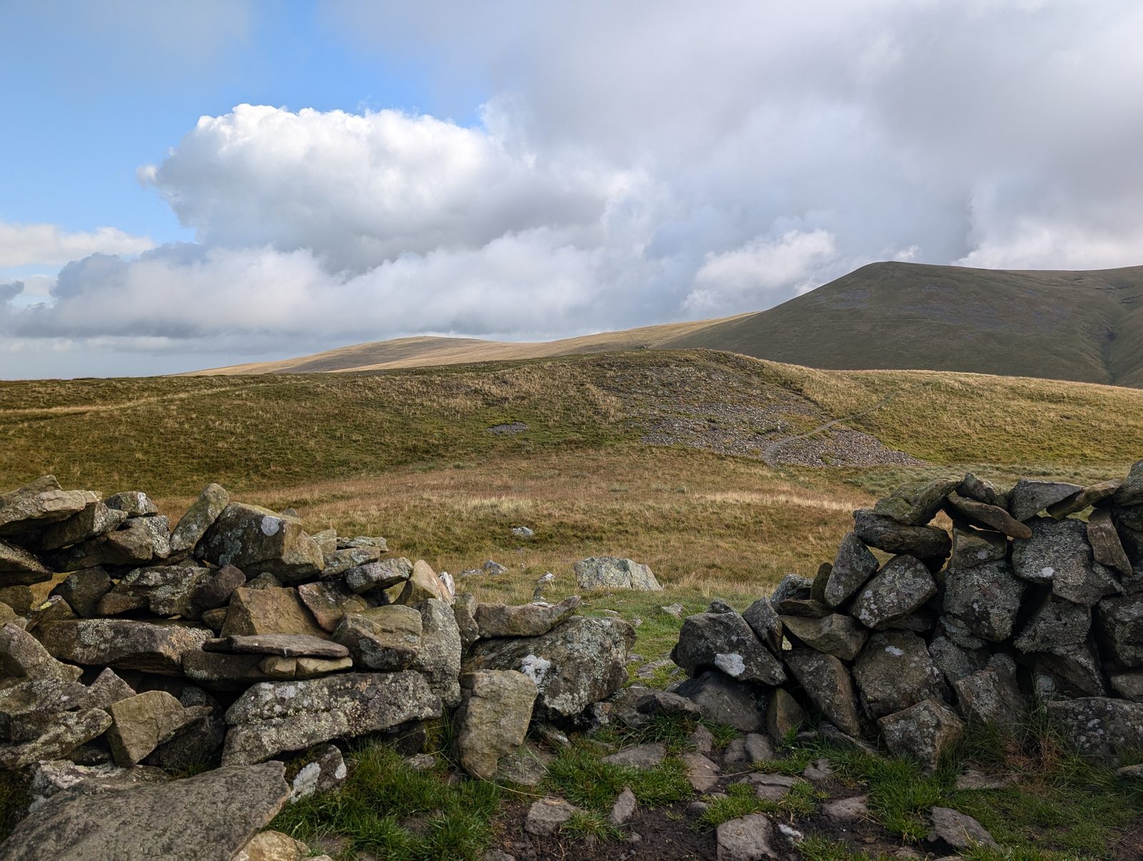 A gap in a drystone wall looking across a broad col of pale moor grass to a scree-scarred green fell rising beyond