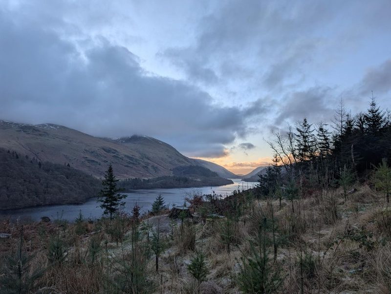 Thirlmere lying grey in its valley at dusk, with young conifers and frosted grass in the foreground and a band of orange light on the horizon