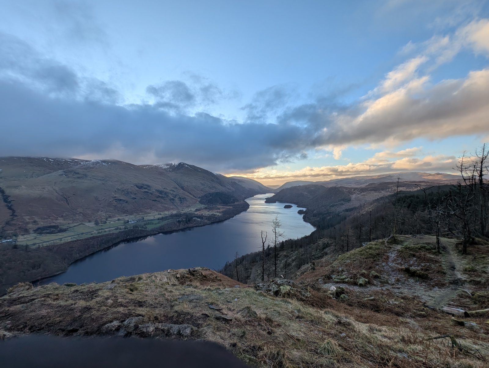 Thirlmere from high above, its full length shining between forested slopes, with dead standing trees on the crag edge in the foreground