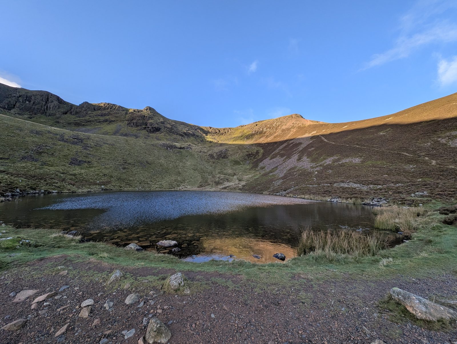 Bleaberry Tarn in its corrie, the dark water ringed by grass and stones, with crags and a sunlit top on the ridge above