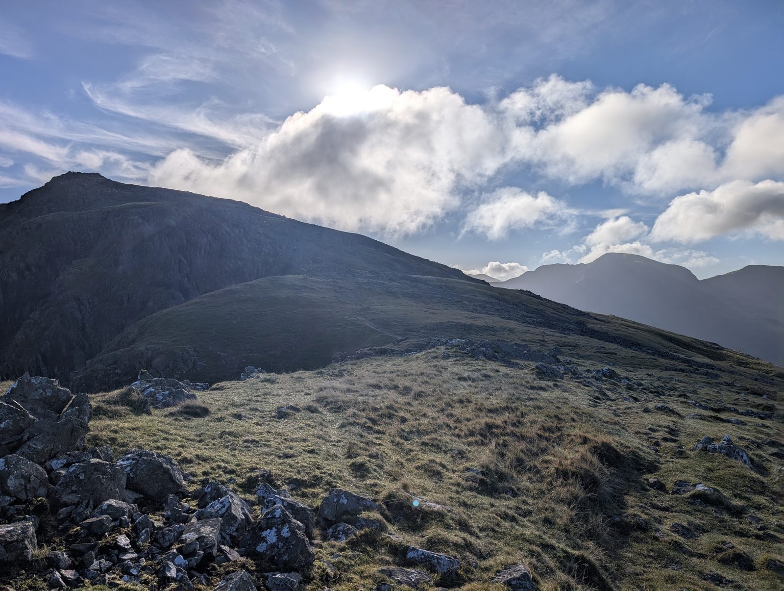 The dark bulk of High Stile ahead across grass and broken rock, the sun behind bright cloud above it