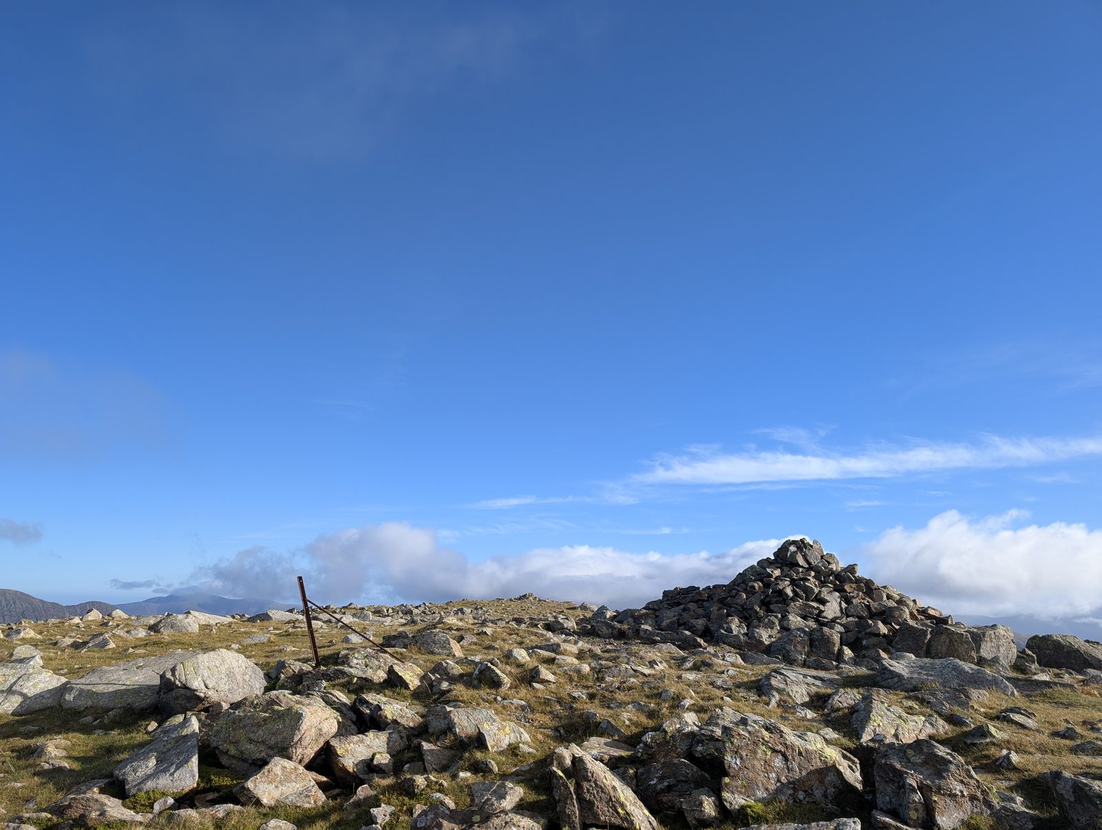 A large cairn and an old iron fence post on the bouldery eastern top of High Stile, far fells on the skyline and white cloud low behind