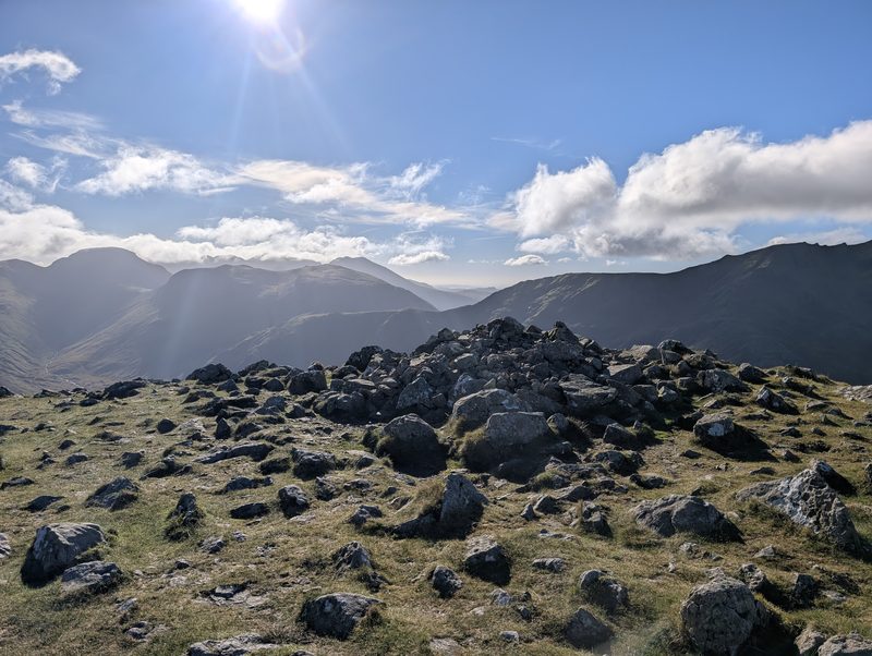 The rocky top of High Crag, a heap of grey boulders in grass, with fells and a deep valley beyond