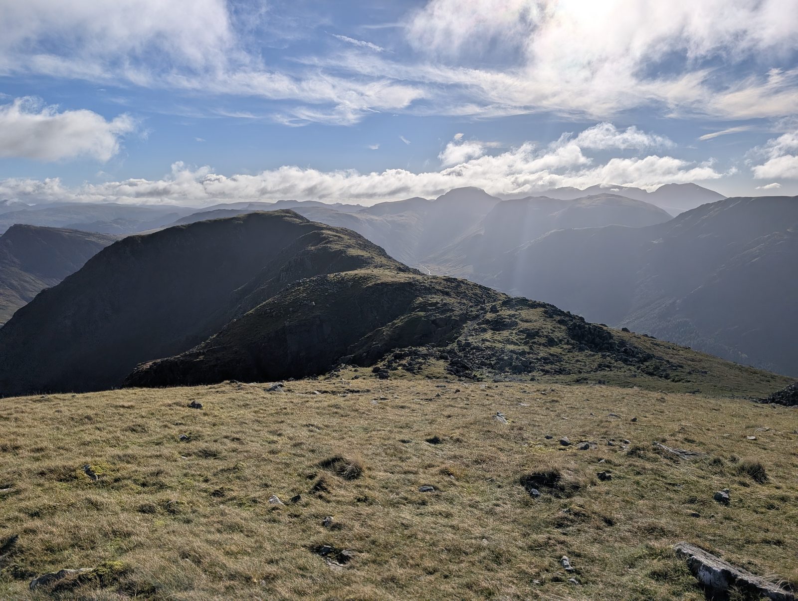 The grassy ridge running on east, a dark, crag-edged top ahead and further fells hazy in the sun