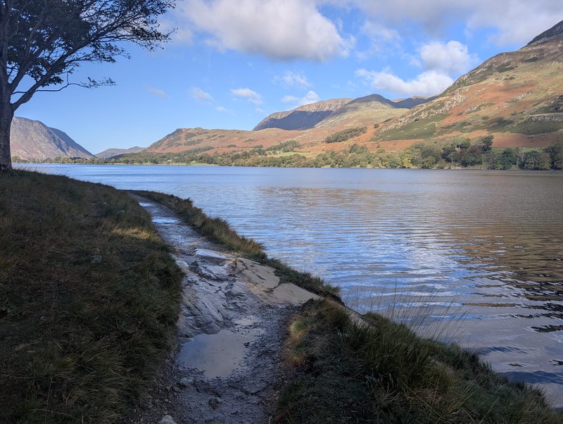 The path along the edge of Buttermere, wet rock and grass beside the rippled water, a tree overhead and bracken-brown fells across the lake